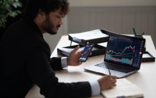 Smart Focused Indian Broker Investor, Stock Trader Using Smartphone And Laptop To Analyze And Invert In The Financial Market Of Cryptocurrencies, Buy Or Sell Crypto Coins, Planning Strategy