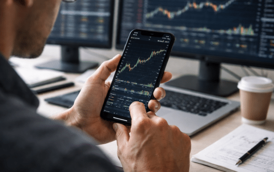 A Trader Analyzes A Financial Price Chart On A Smartphone While Multiple Market Charts Display On Monitors In The Background.